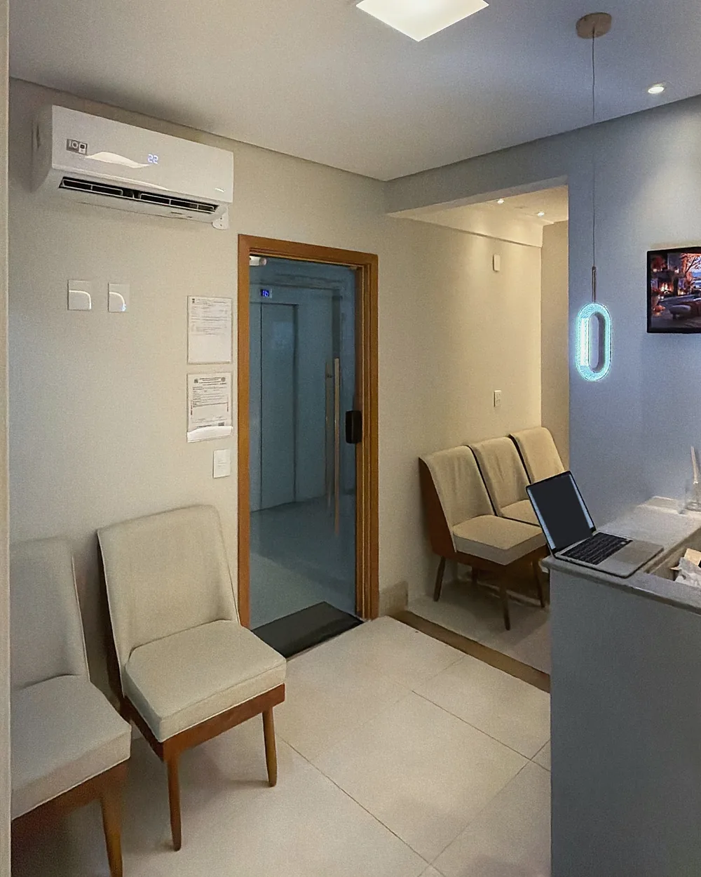 Sala de espera com poltronas claras, porta de vidro e luminária oval